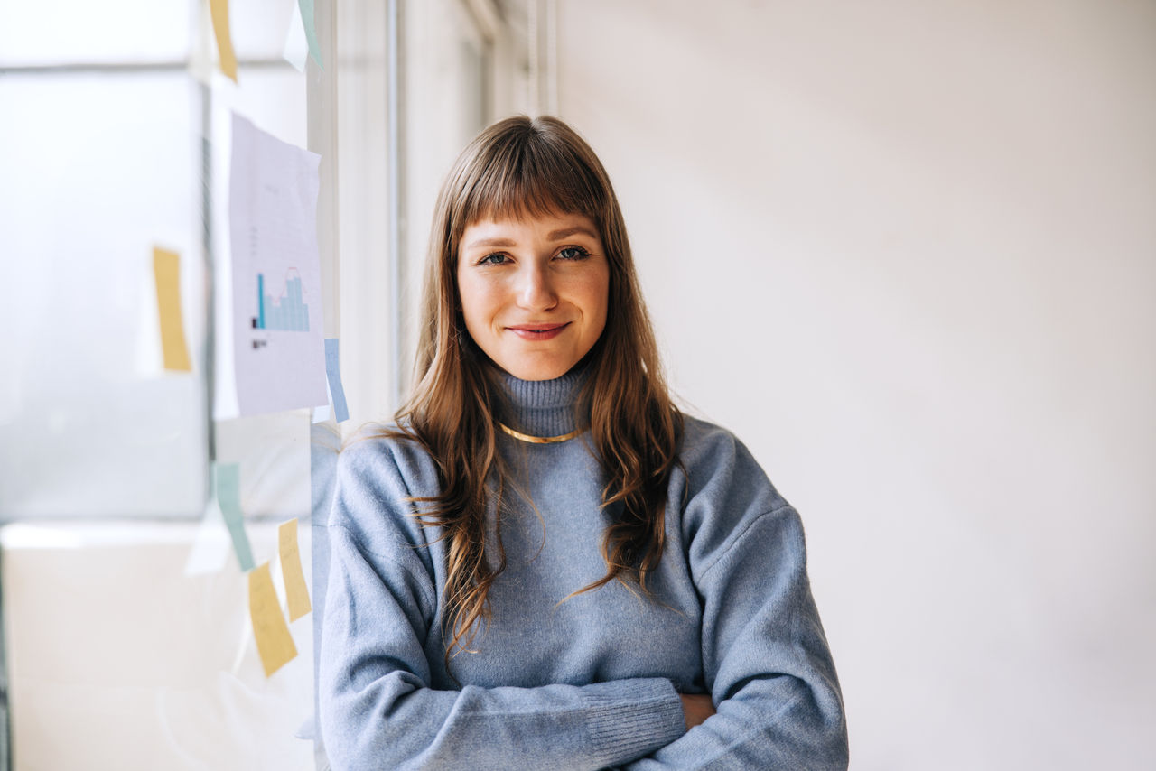 Portrait of a young businesswoman looking at the camera while standing next to a glass wall in an office. Happy female entrepreneur organising her creative ideas using sticky notes.